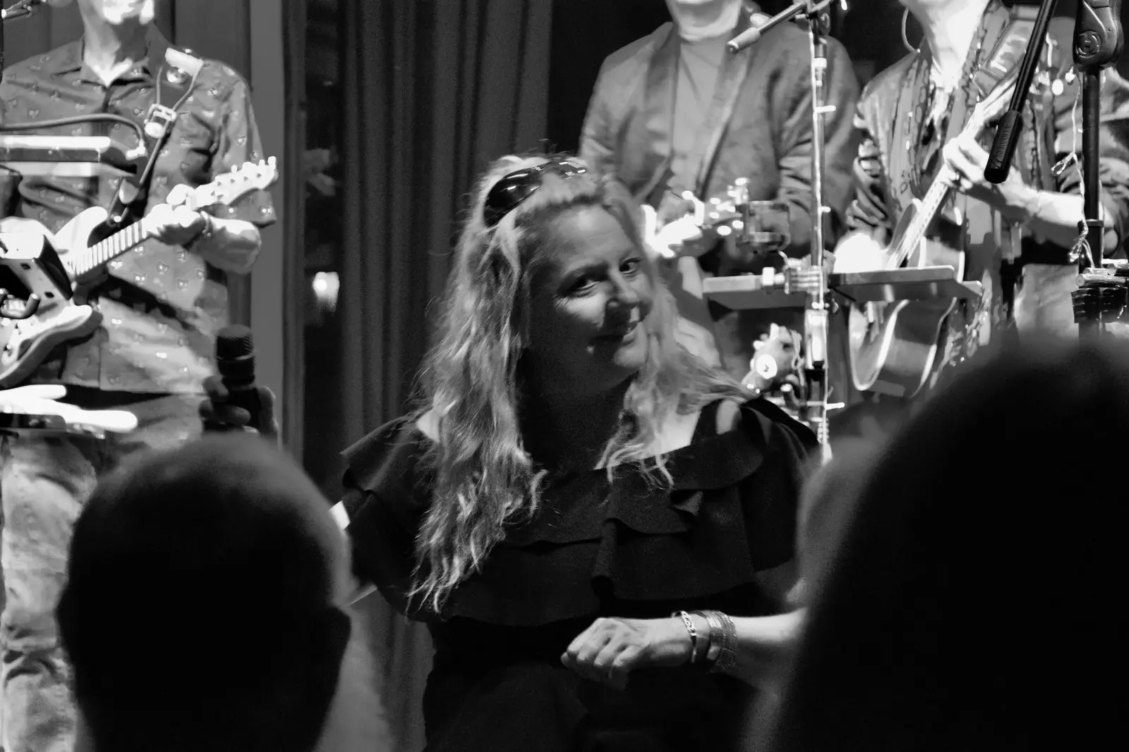 Photographed over the heads of the audience, a singer smiles between songs while the rest of the band play behind her.